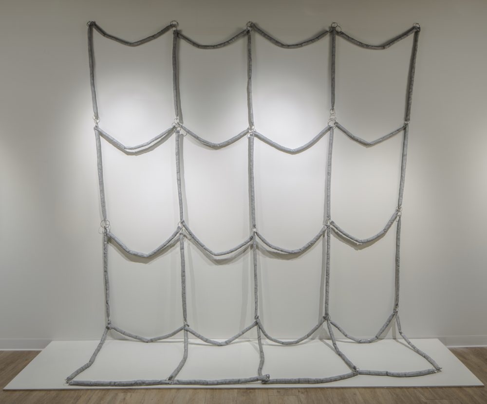 Soft abstract grid sculpture hanging on a wall and resting on the floor.