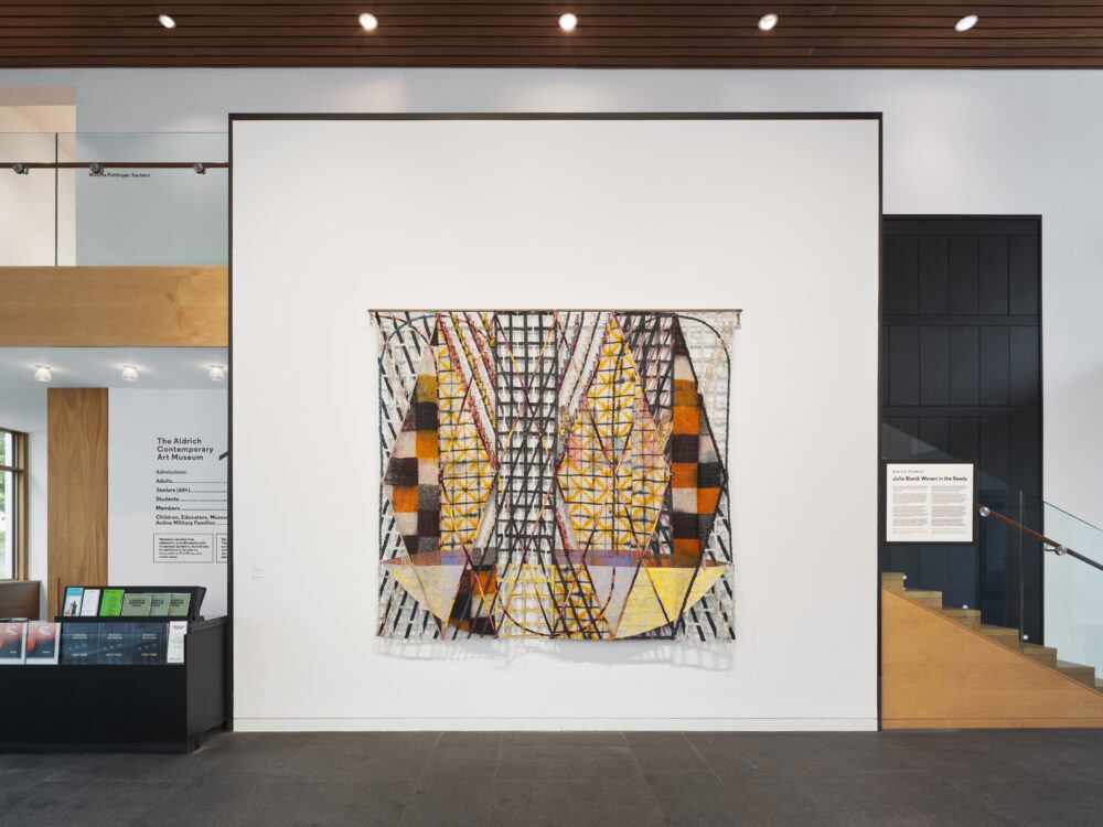 Colorful, Dyed, Woven, Braided, Tied, Hand-Sewn Monumental Tapestry hung on a white wall in a museum.