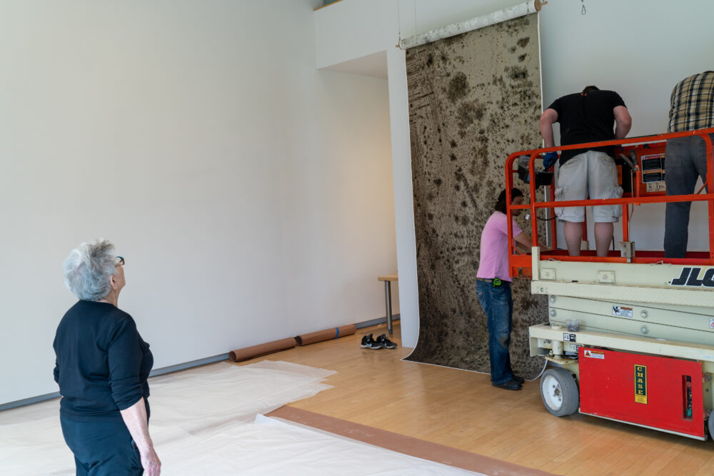 Person watching three installers on a scissor lift installing a large-scale art work on a white wall.