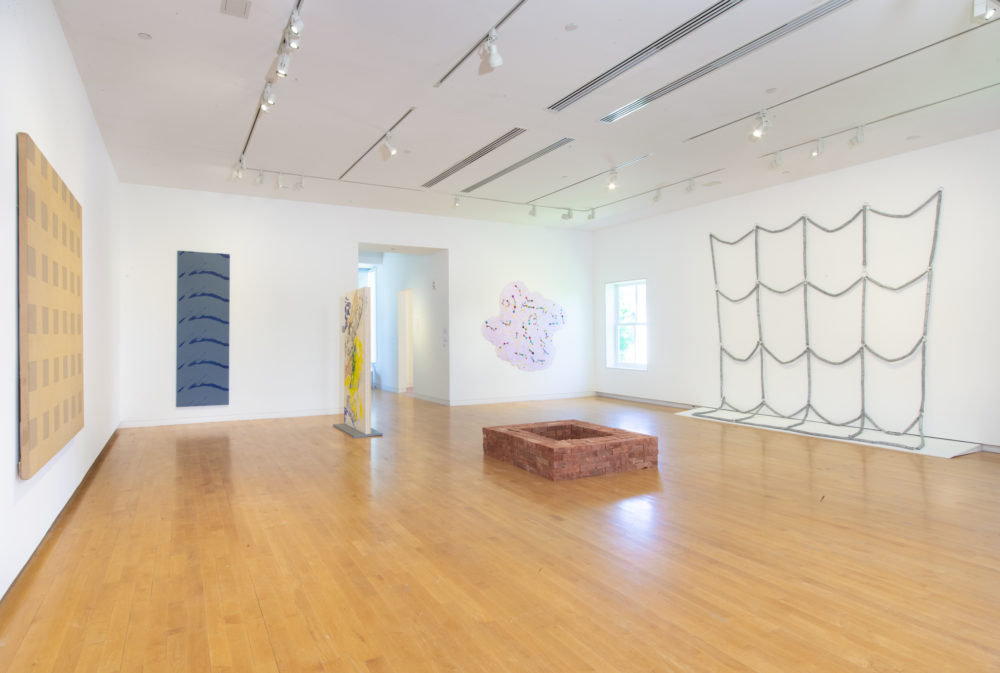 Gallery with painting and wall mounted sculpture and a freestanding square brick sculpture in the center of the room.