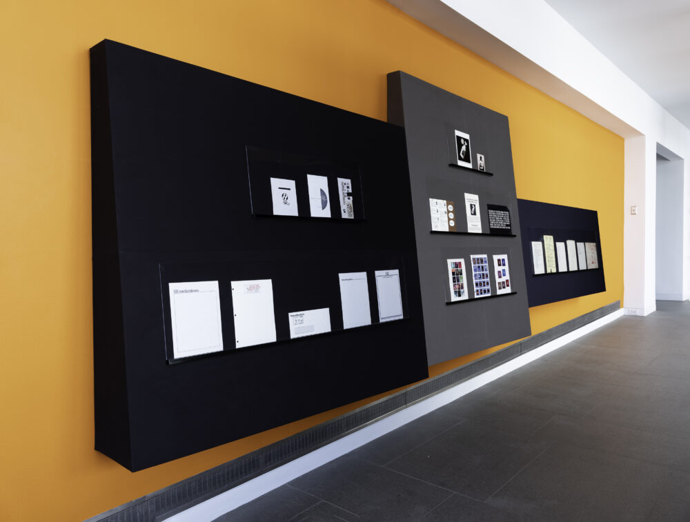 Archival Branding Materials from Syntonic Research displayed on custom black shelving that is hung on an orange wall.
