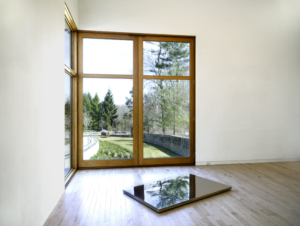 Mirrored stainless steel rectangular flat floor sculpture in front of a window in the Museum gallery.