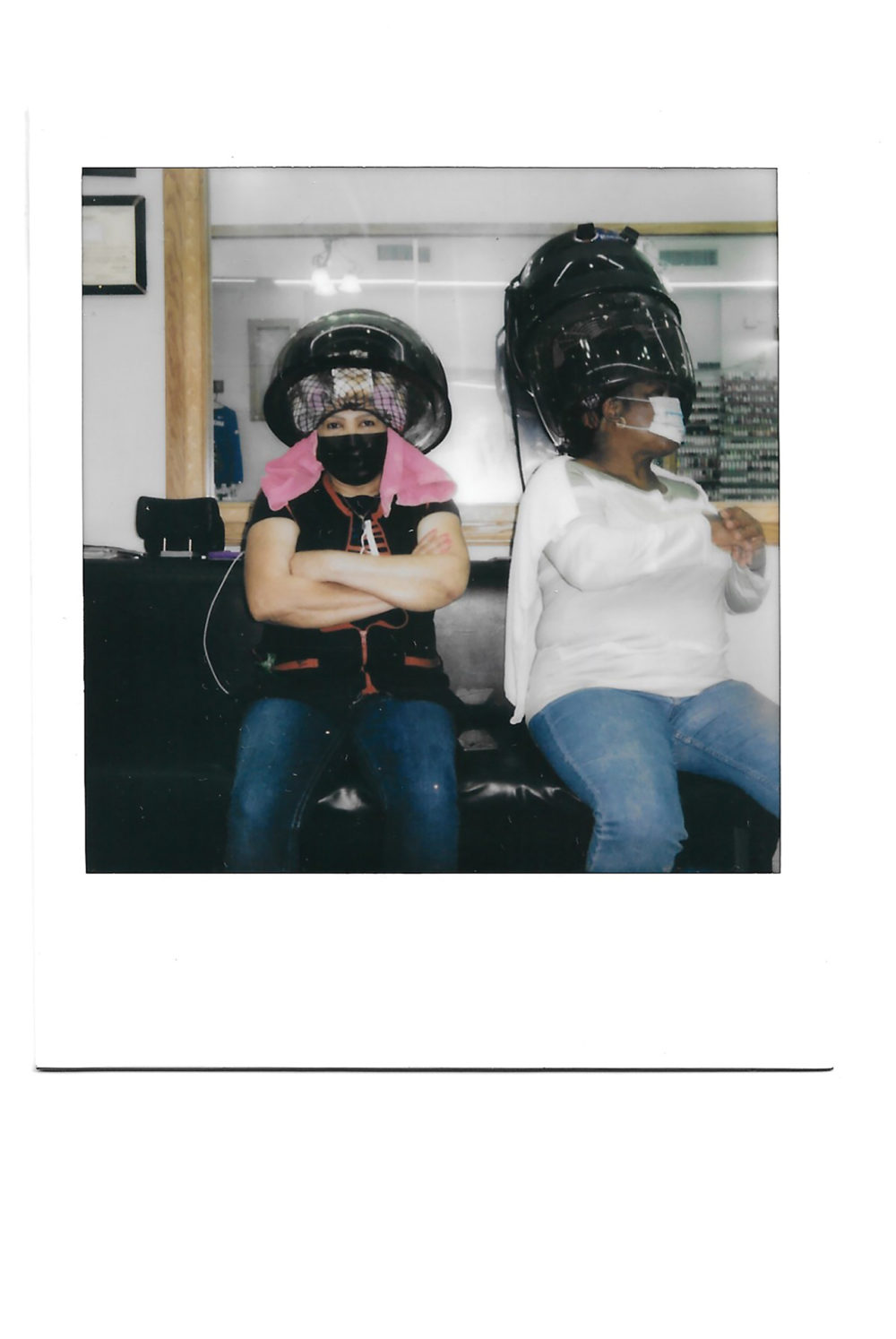 Two people sit in a salon underneath a hair dryer