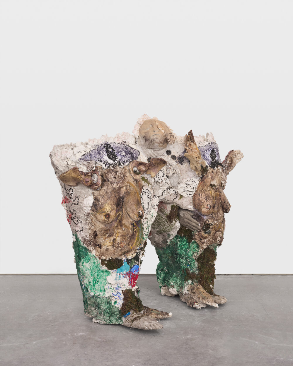 A green and brown sculptural art work of an abstract creature with two pregnant human torsos, a face, and hands and feet.