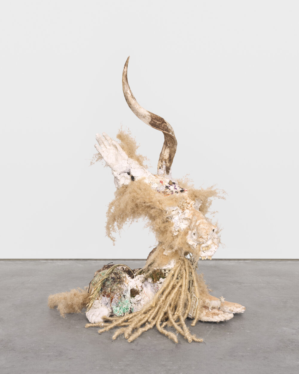 Beige colored sculptural artwork with a spiral horn atop, a hand, feet, and many hair-like textures.