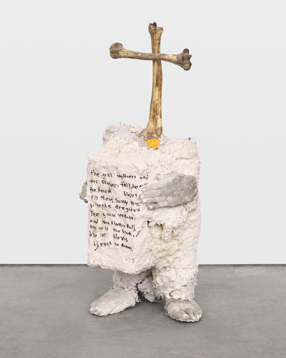 Sculptural white artwork with hands and feet on an abstract figure with scripted writing on its front side with a bone cross atop.