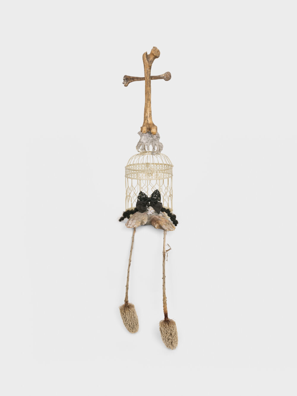 A sculptural art work with a white birdcage, gold bone cross, feet, and a black bow.