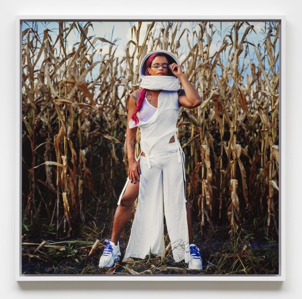 Photograph of a person wearing sneaker anda helmet-like headpiece, with a long fuchsia braid standing in a cornfield.