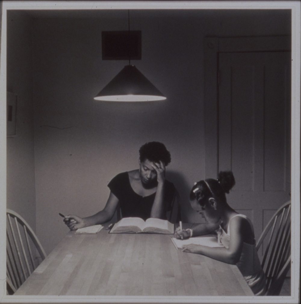 A black and white photograph of two individuals sitting at a table reading and writing