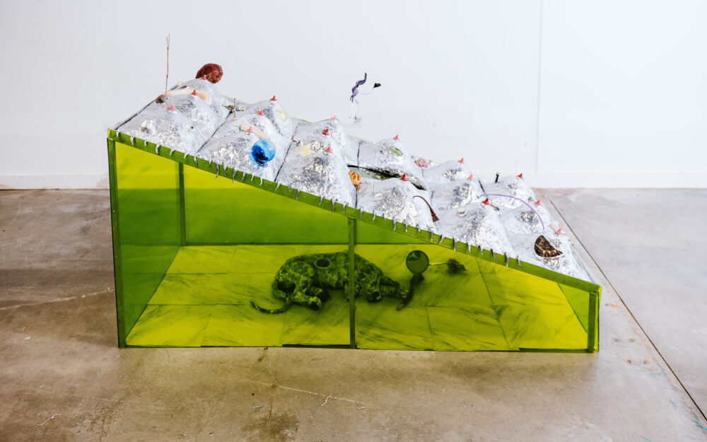 A sloping sculpture on the floor with transparent chartreuse sides revealing a dog-like figure and a silver mounded top.