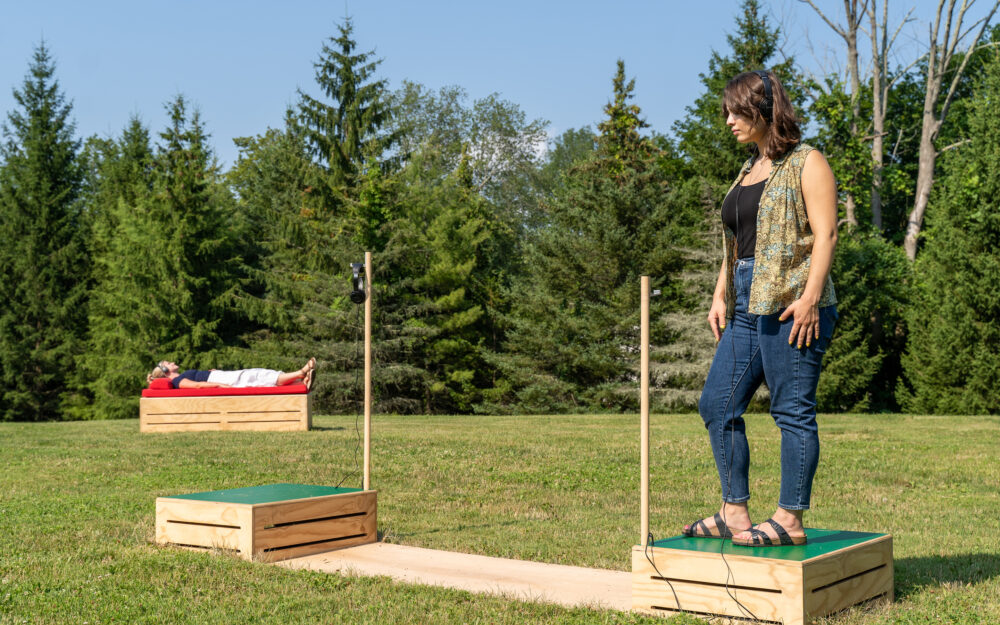 A woman wearing headphones and standing on a soapbox in the foreground and a reclining woman on a soapbox wearing headphones in the background.