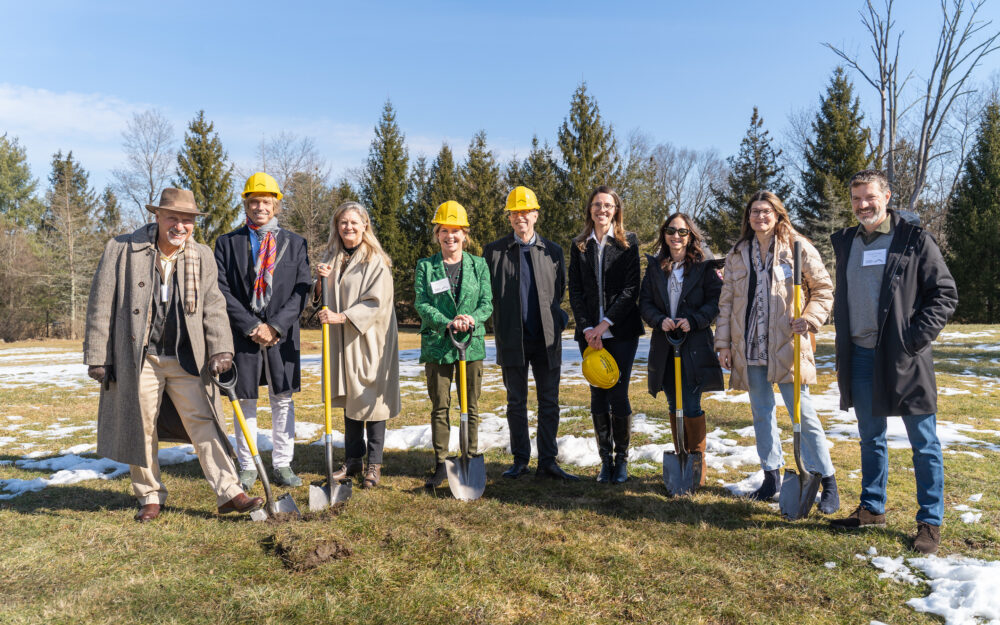 A group of people holding shovels and wearing hard hats in a line outdoors on the grass.