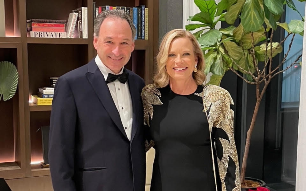 A man in woman in black tie attire in front of a bookshelf and plant.