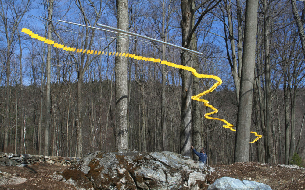 Multi-part suspended sculpture of yellow squares strung together forming a curving line through a winter forest canopy.