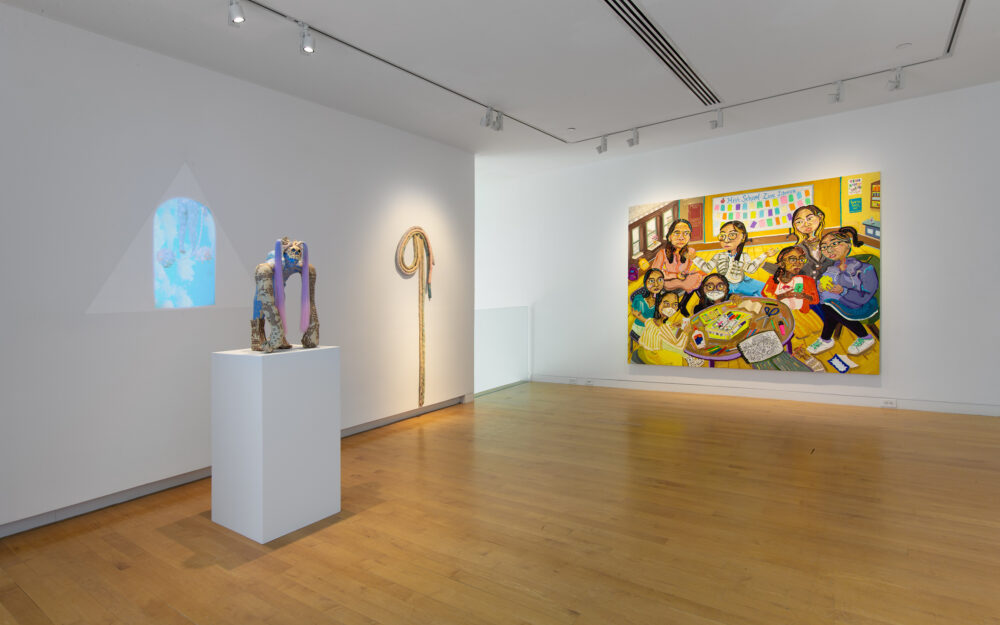 Install view of a gallery with a figurative sculpture to the left with a projection in the background, a soft sculpture hung on the wall at center and painting.