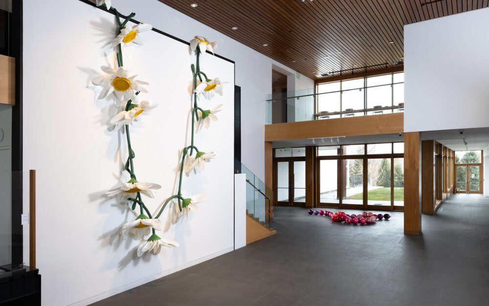 large scale sculpture of a daisy chain made of gardening materials hung on a wall with a large string of peals made of pink bowling balls coiled on the ground
