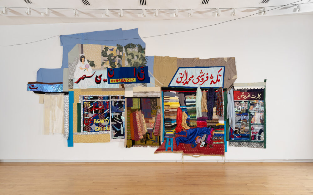 Large-scale wall hanging textile work depicting a bazaar stalls selling bolts of fabric, a beauty salon, and more.