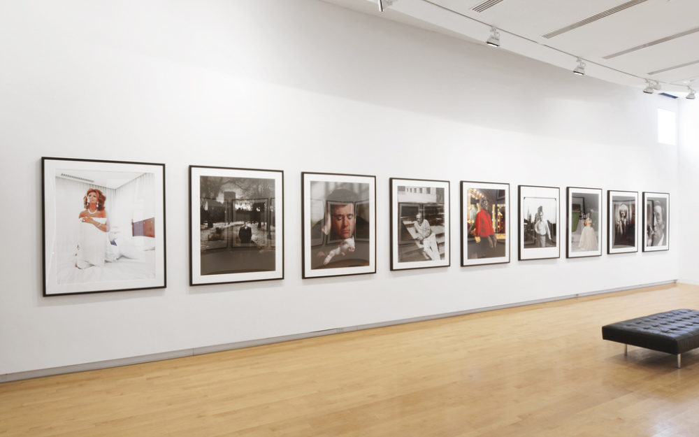 Nine square portraits hang on gallery walls in black frames