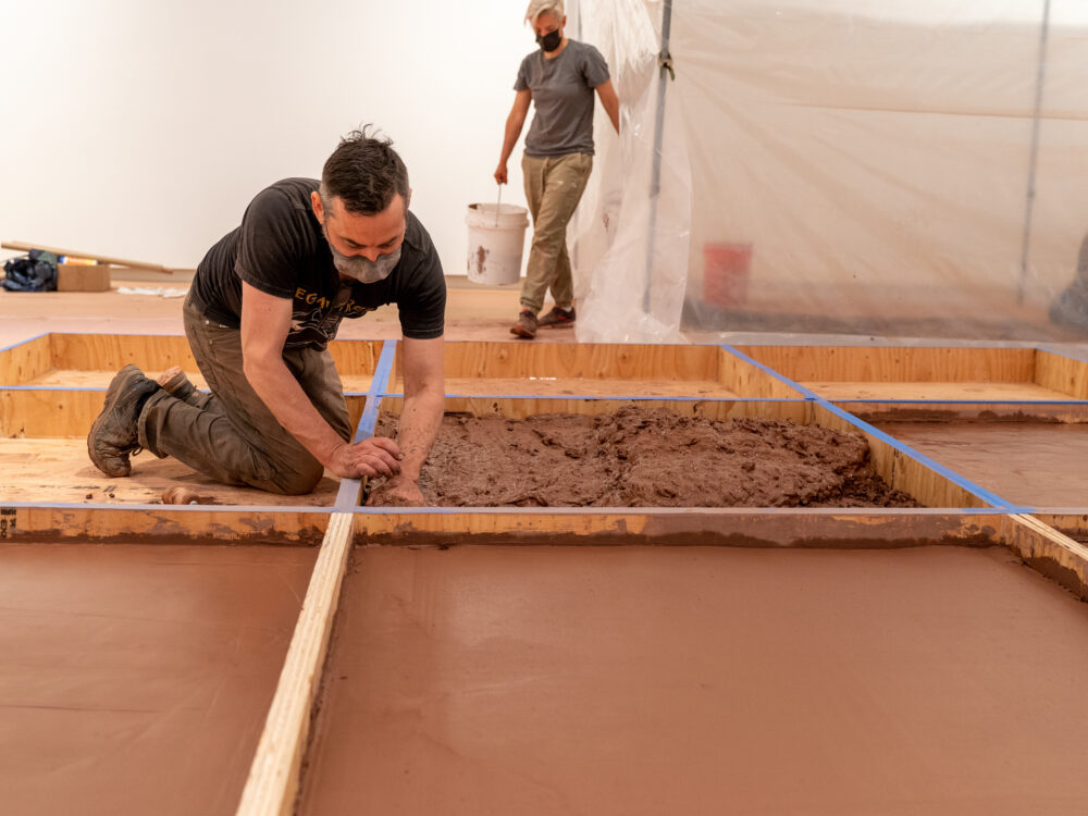 Person pressing wet red clay into a wooden gridded armature.