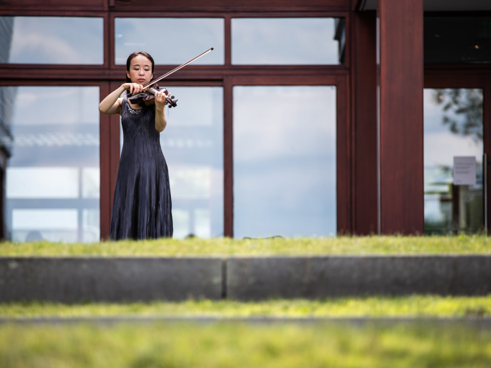 A violinist performs with large windows in the background