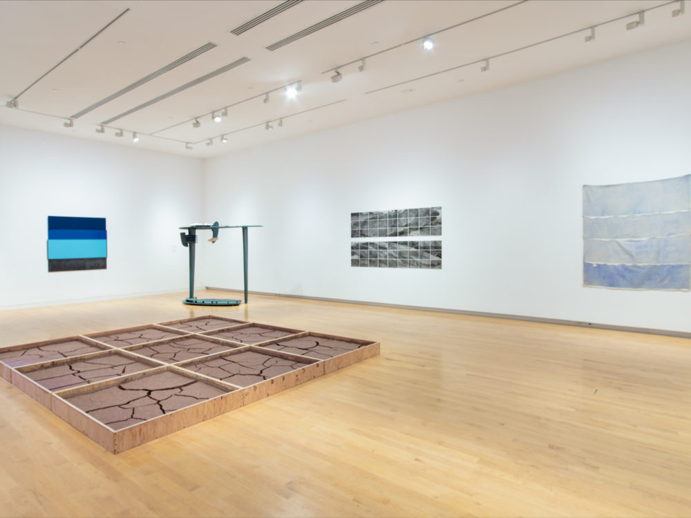 Gallery with paintings on the wall and two abstract sculptures, one tall and made of dark metal, the other on the floor, a wooden grid filled with red clay.