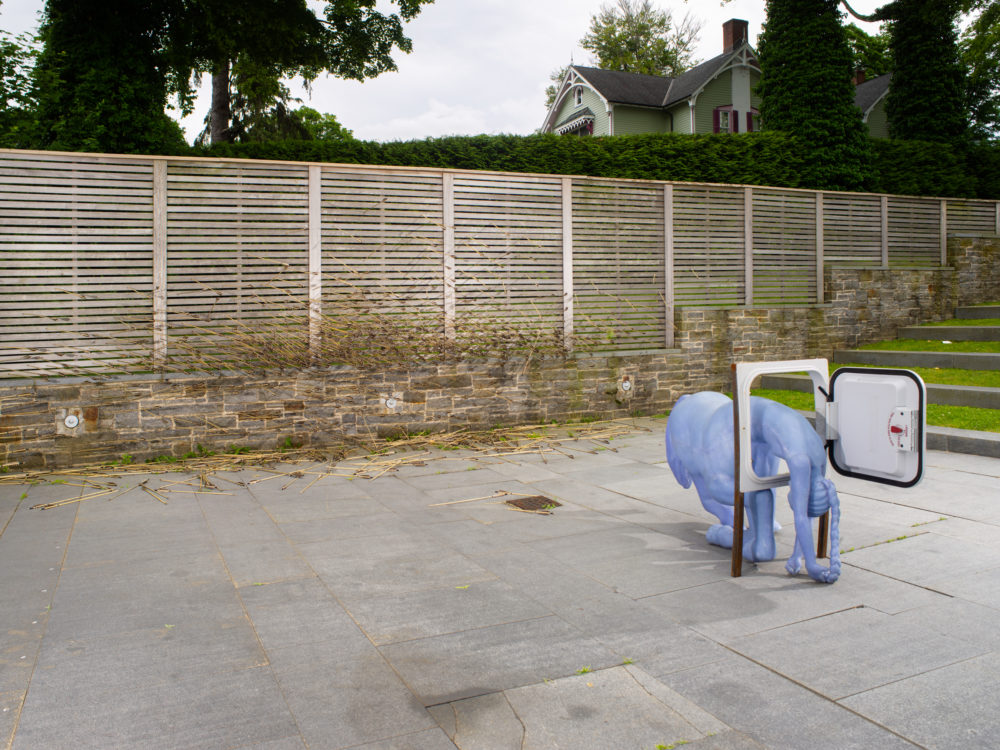 Outdoor sculpture of a blue centaur kneeling down though a school bus escape hatch with many arrows on the ground and stuck in the fence in the background.