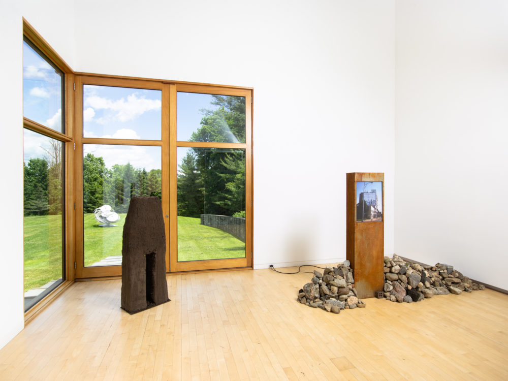 Two freestanding sculptural works, one with a pile of stones, the other made of earth. Through the window there's a silver sculpture outside.