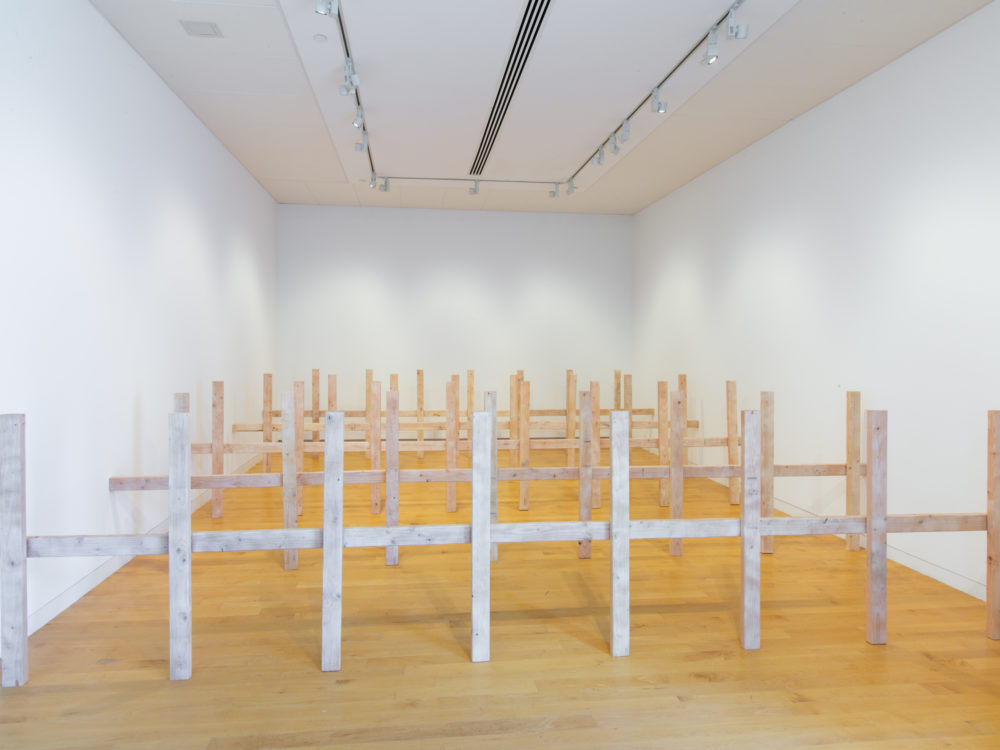 Several unpainted wooden fences in a row that fill a gallery space.