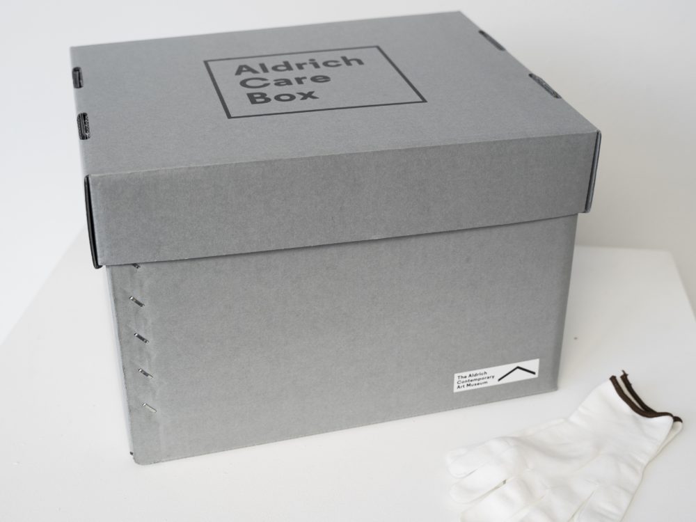 Gray archival box with "Aldrich Care Box" on the lid in black lettering with a pair of white gloves.