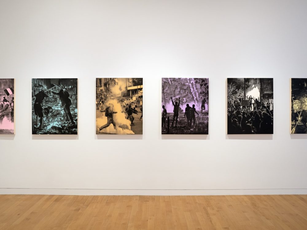 Six single color and black paintings of protest scenes hung in a line on a white wall.