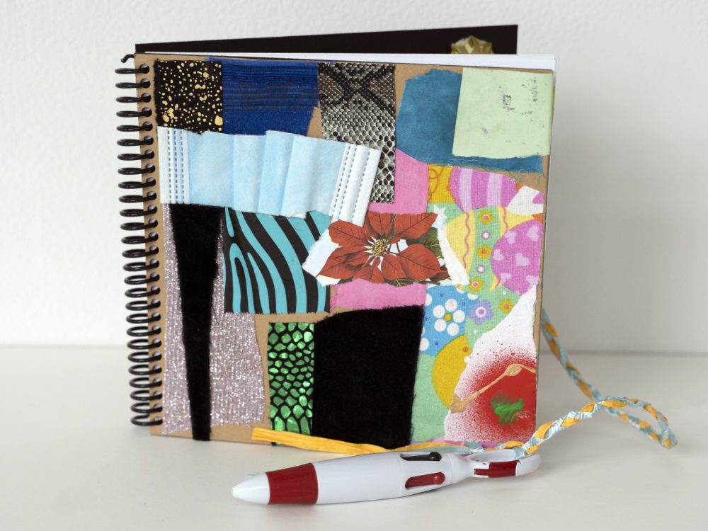 Image of an upright spiral bound notebook with collaged elements on the cover including a piece of a disposable face mask and shiny fabrics. A pen is attached.