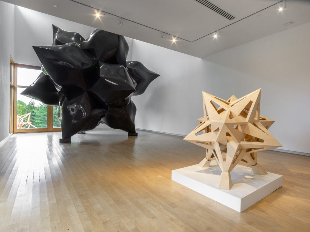 Smaller wooden star sculpture in foreground, large-scale black star sculpture in background.