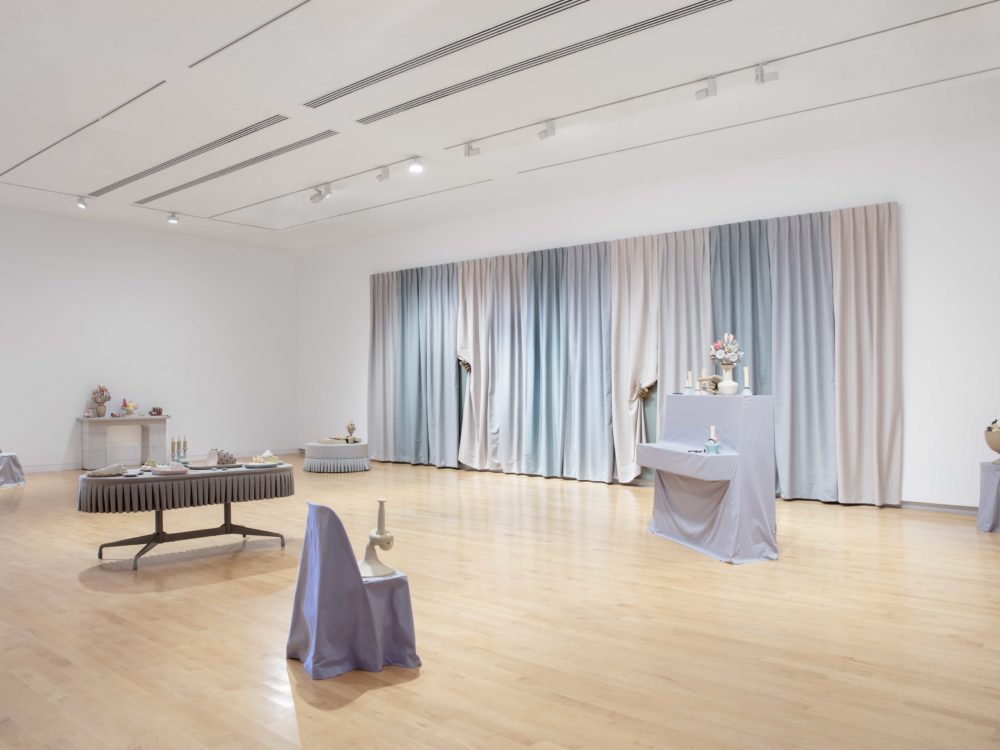 Overall installation view with gray slipcovered furnishings with surrealist clay objects and a large-scale multi-colored curtain on the back wall.
