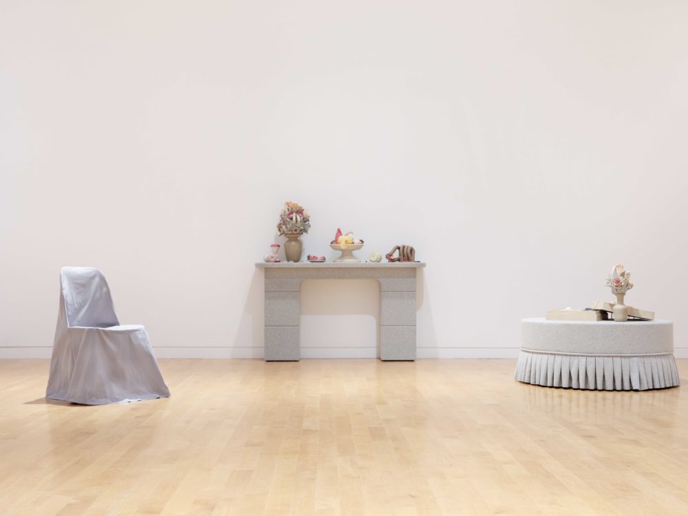 Gray slipcovered chair to the right, gray mantle with bouquet and fruit bowl at center, gray pouf with bouquet at right.