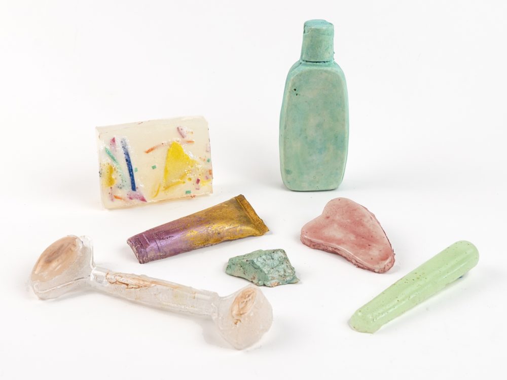 Cast sculpture colored objects related to self-care practices including a bar of soap, facial roller, gua sha stone, and tube and bottle of lotion.
