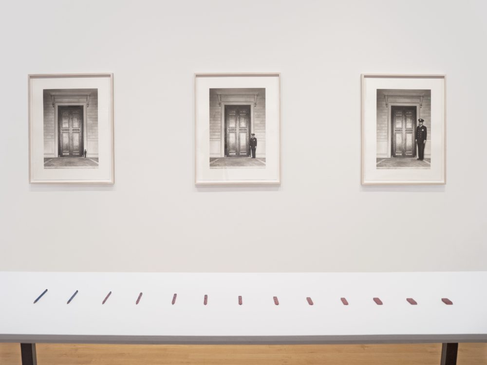 Installation view with a table in the foreground with small sculptures of a pencil turning into an eraser; on the wall behind are three framed drawings.