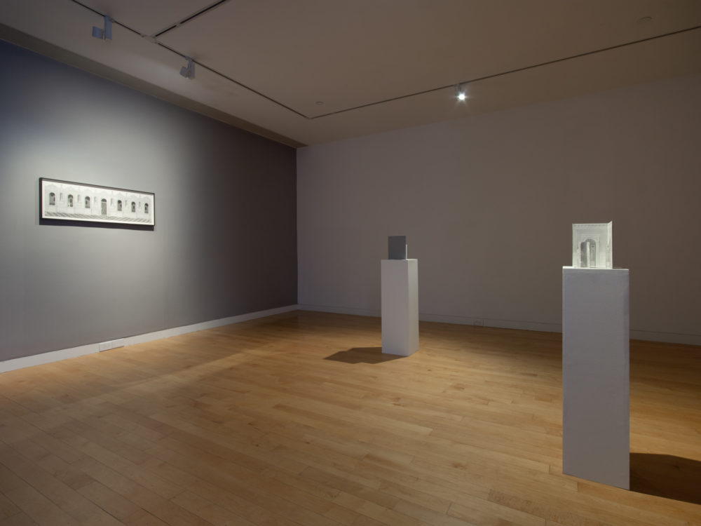 Gallery with gray walls and a framed drawing hung to the left and two pedestals with three-dimensional drawings placed on top.