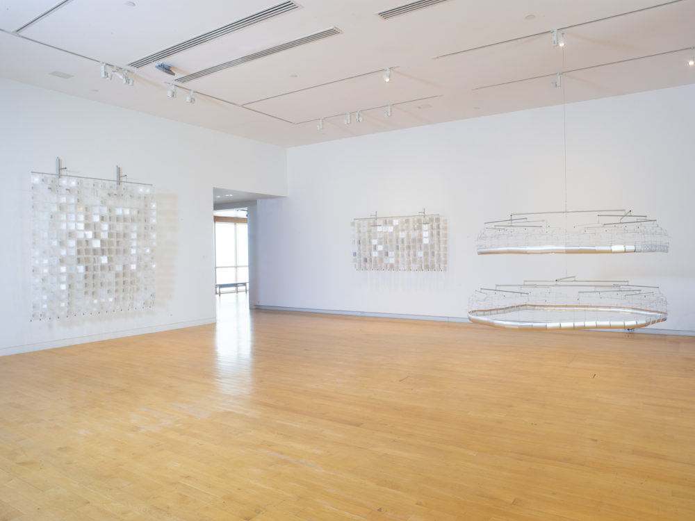 Gallery space with three sculptures made of reflective material and wire, two are hung on the wall and one is suspended from the ceiling.