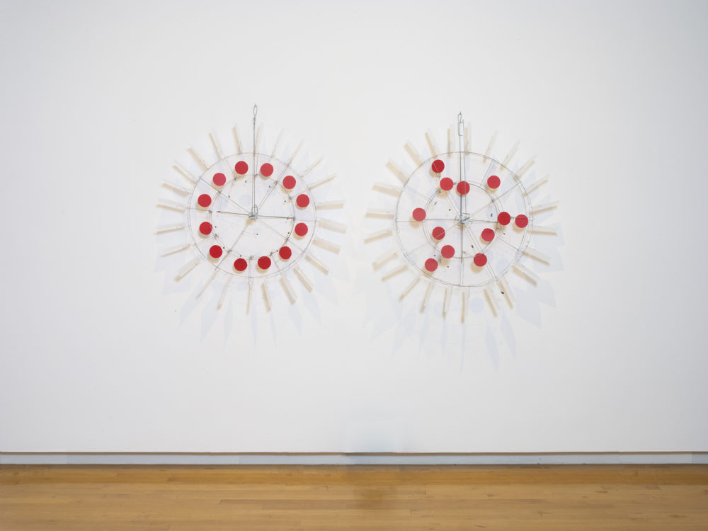 Two circular sculptures hung on a wall with red dots in the middle and reflective material outer pieces connected by wire.