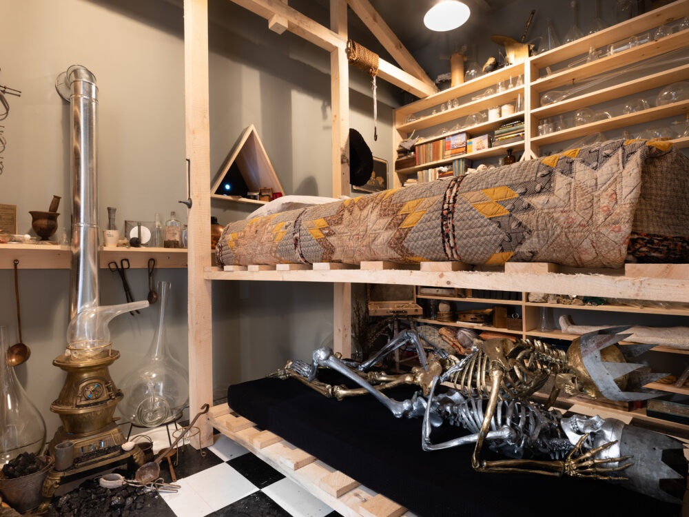 A closeup shot of a small room with numerous alchemical artifacts. A bed sits in the center with silver and gold life-size skeletons embracing underneath.