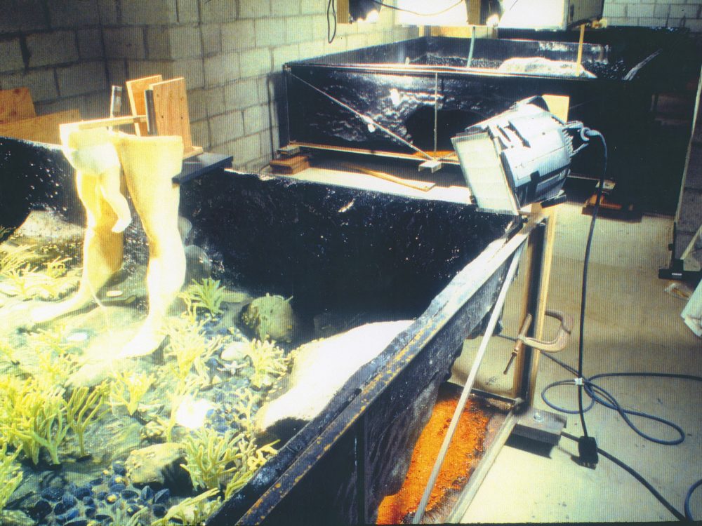 A large square frame filled with a false ocean floor with two wax legs, one a man's the other a baby, standing on the ground