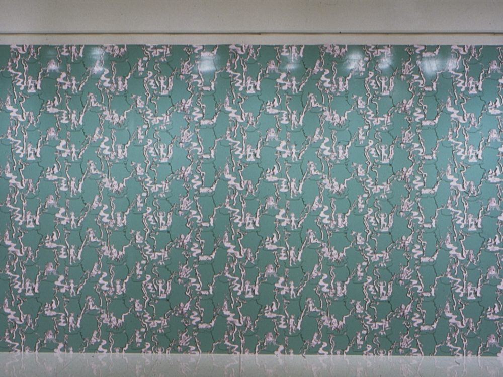 This is a photograph of a work by Mary Esch which looks like green wallpaper which many little white drawings repeated along the wall